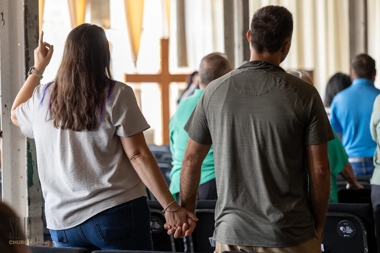A couple holding hands in worship with the cross in the window light