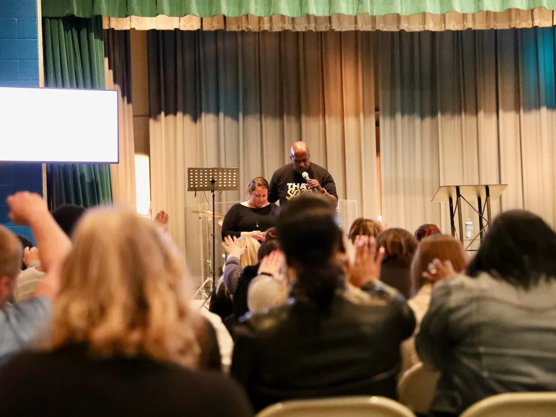 Preaching from the cafeteria stage with hands raised in the room